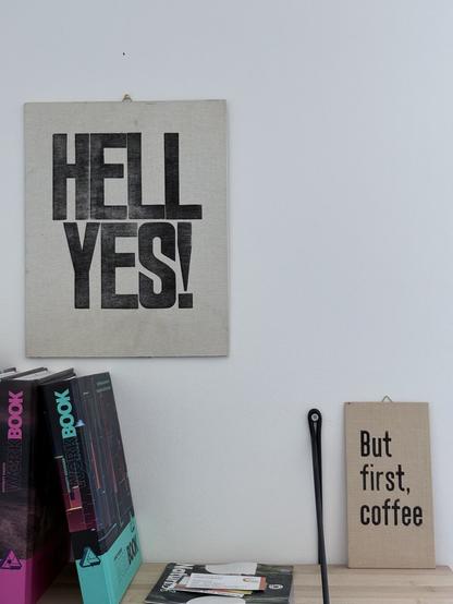 A sign above a book shelf reads „HELL YES!“. On said book shelf, though, a smaller sign rests against the wall. It says „But first, coffee“.