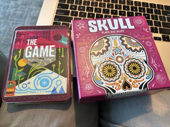 Two board game boxes. A small metal case with the card game "The Game" on the left, and a square box with the game "Skull" on the right.