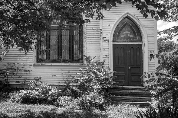 Black and white image of the front door and stained glass windows of an 1898 African Methodist Episcopal Church building with white peeling paint and overgrown landscaping.