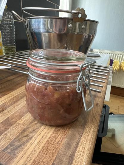 A mason style glass jar with a darkish chutney inside sits on a kitchen counter. There is a large stainless steel jam pan sits on the cooker behind.
