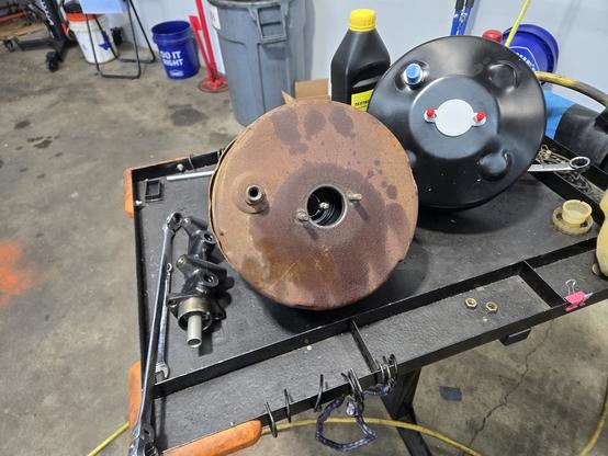 An image of two brake boosters on a table. One is old and rusty, the other is new and painted black. There is a new master cylinder next to the old booster.