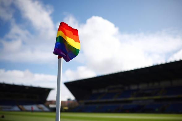 Im Vordergrund weht eine Pride-Eckfahne, im Hintergrund in der Unschärfe die Tribünen eines Stadions. (Credit: IMAGO/Shutterstock)