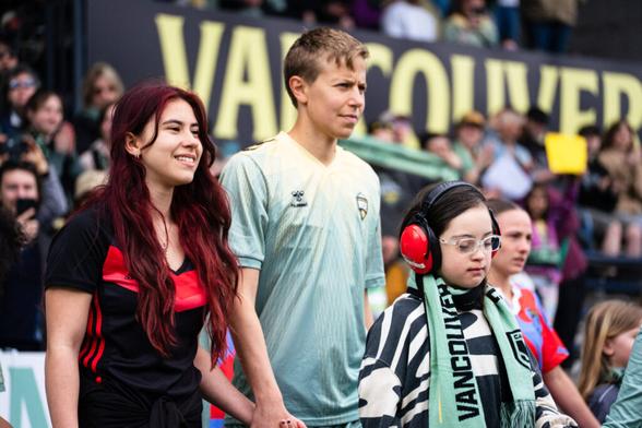 Quinn, eine fußballspielende Person, läuft ins Stadion ein, an ihrer Hand eine Einlauf-Jugendliche mit langen, offenen Haaren, vor ihr versunken ein Einlaufmädchen, im Hintergrund in der Unschärfe die Tribüne mit Zuschauenden. Credit: IMAGO/Sports Press Photo