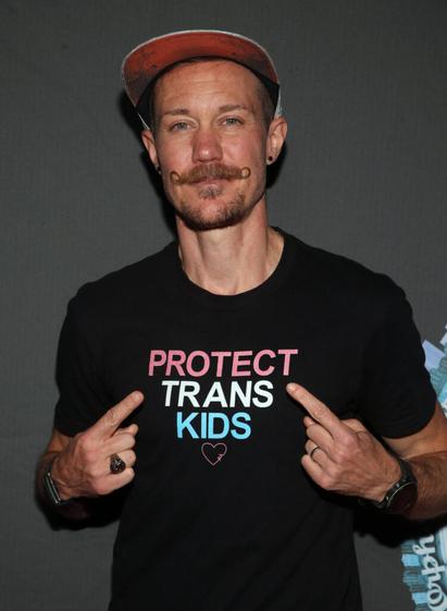 Chris Mosier mit einem Shirt, auf dem steht: "Protect Trans Kids", darunter ein Herz, er zeigt mit den Zeigefingern beider Hände auf die Schrift. Credit: IMAGO/ZUMA Press