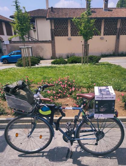 La mia bici da città blu vista da di fianco, parcheggiata davanti ad un'aiuola fiorita. La bici ha una borsa appoggiata sul portapacchi anteriore ed uno scatolone su quello posteriore. Dietro si vede una cascina