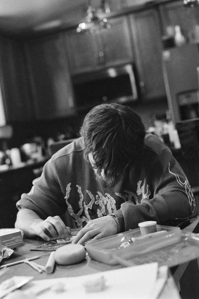 A black and white photo of a teenage boy sitting at a kitchen table, deeply focused on shaping something with his hands. His long hair partially hides his face as he leans over his work. The table is cluttered with small tools, a round piece of clay, and crafting supplies. The background shows a lived-in kitchen with wooden cabinets and soft lighting