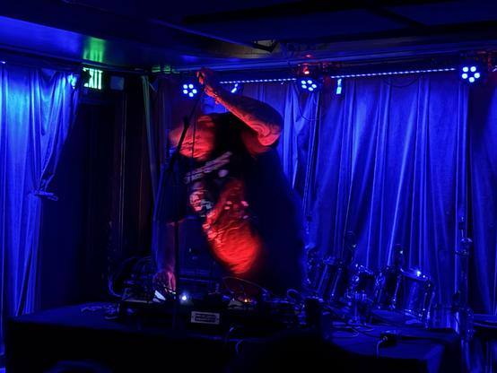 Very large man poised dramatically over a deck of crazy electronic noise making gear. Uplit in red in a purple room.