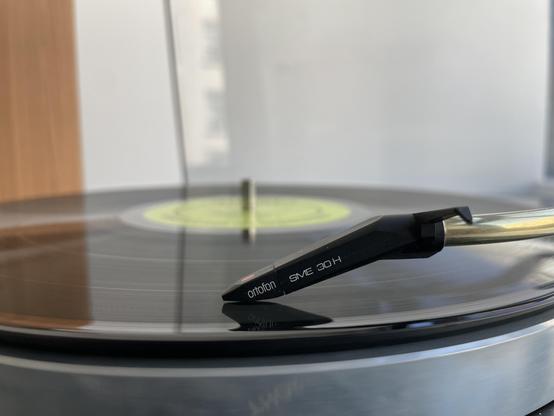 A close-up of a turntable needle resting on a spinning vinyl record, with wooden cabinetry in the background. The needle is labeled "ortofon SME 30 H."