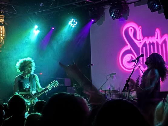 Three-piece heavy metal band on stage lit strongly green on the left, pink neon style logo on the right, dramatic contrasts.