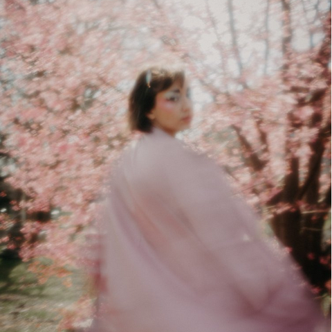 A femme, genderqueer person stands among cherry blossom trees. They are wearing a pink jacket or cloak with their back to the camera. They look over their shoulder. Their hair is in a short brown bob and they are wearing light blue eyeshadow. The camera is in soft focus.