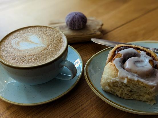 Colour photograph of a cup of milky coffee and a cinnamon bun with icing on top, both served on duck egg blue speckled crockery. There is a small sea urchin stuck to a bit of driftwood behind them on the table top.
