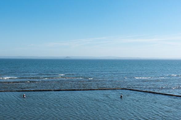 Colour photograph of a stretch of blue sea. A section at the front is surrounded by a stone wall, forming an outdoor swimming pool. The water in this area is still, and the heads of two swimmers are visible. The sea beyond has small white crested waves. The sky is blue.