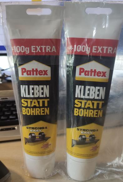 Bild von zwei auf dem kopf bzw dem deckel stehenden klebstofftuben 'pattex kleben statt boren' in einer durchsichtigen plastiktüte, auf einem schreibtisch stehend. Im hintergrund sind noch eine tastatur und zwei bildschirme auszmachen, der bildausschnitt stellt die klebstofftuben prominent mittig und sehr groß dar.