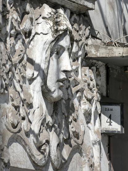 Decorative relief on a tenement house wall — face of a young woman, surrounded by leaves. She was sculpted en face, but the photo is made in such angle we see her profile.
Also, a house number (in a form of small lantern), 4 Antoniego Józefczaka street.
