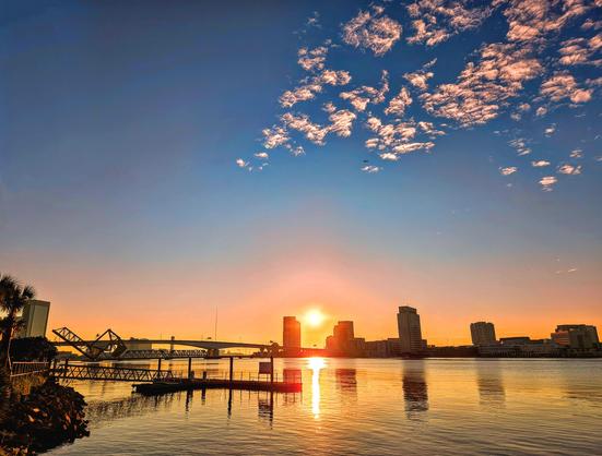 Morning view across the Saint Johns River towards the downtown Jacksonville skyline and bridges as the sky changes with sunrise colors rising as the night lights and skyline silhouettes emerge from the darkness. A bright golden sun creates a sunburst above the bridges,  casting reflections upon the wster below and tiny pop-corn like clouds above.