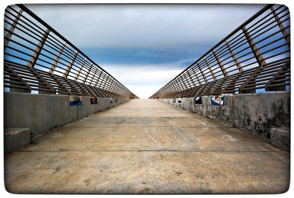 A view of a pier that pokes out into San Francisco Bay from the Embarcadero.  The view is from the ground looking forward and up at the pier's angled bracket < > railings as they recede, symmetrically, into the distance. There are cloudy blue skies above and yellowing concrete covering the deck of the pier.

For those wondering, the lens is a Sigma 8-16mm ultrawide lens... on an old crop sensor Pentax body.