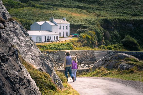 A scenic photograph taken on Cape Clear Island, County Cork, showing two people walking arm in arm along a winding path bordered by rugged rocks and grassy slopes, with white cottages nestled in lush green hills in the background under soft daylight.