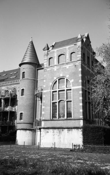 Black and white photo of a historic brick factory building with large arched windows and a cylindrical turret topped with a conical roof, evoking a castle-like appearance. The structure is set against a clear sky and surrounded by a grassy area with small flowers and a low fence in the foreground.