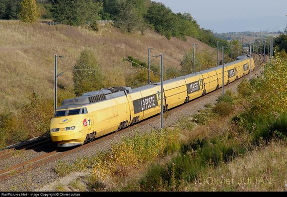 Photo d'Olivier Julian. Plan large montrant l'ancien TGV postal sur ses rails en pleine nature. Il y a une composition classique à 8 wagons et 2 motrices. Une voiture sur deux a le logo de La Poste en blanc sur fond gris. Le reste est peint en jaune caractéristique de La Poste. Aux extrémités (sur le nez des motrices) est ajouté un logo SNCF.