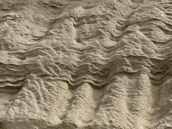 Looking up at a sandstone cliff, all light tan and beige and brown shades. It has been heavily shaped by water with underlying column-like shapes and an overlay of wave-like terraces. These are wavy horizontal ridges that seem to overlap like shingles. 
The light is coming in from above and sideways, highlighting the ridges and casting darker shadowed areas below the wave shapes.
