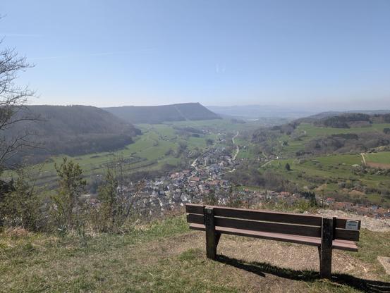 Viewpoint with an empty benchnear the top of Galgenberg (715 m): The valley sprinkled with houses in front, the tree-covered mountain ridge on the left.