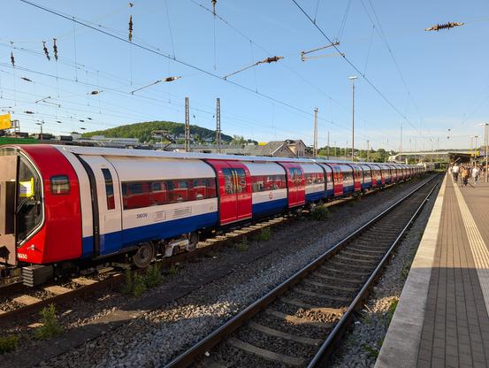 Flache, weiße Wagons mit blauen und roten Details, sowie einem Logo mit der Aufschrift "Underground" stehen auf einem Durchfahrtsgleis an einem Bahnhof. Der Himmel ist blau, die Sonne scheint tief zu stehen und die Wagons werfen lange Schatten nach rechts zum Bahnsteig.