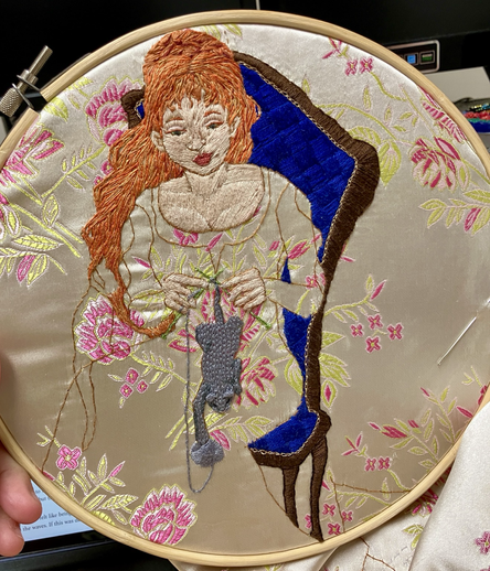 An embroidered picture on a base of cream and pink brocade of a red haired woman in a blue upholstered chair knitting a kitten out of grey yarn.