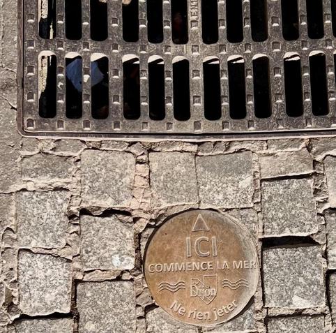 A metal plaque embedded in cobblestone pavement, featuring the phrase "ICI COMMENCE LA MER" (Here Begins the Sea) and "Ne rien jeter" (Do not throw anything), positioned near a metal drainage grate.