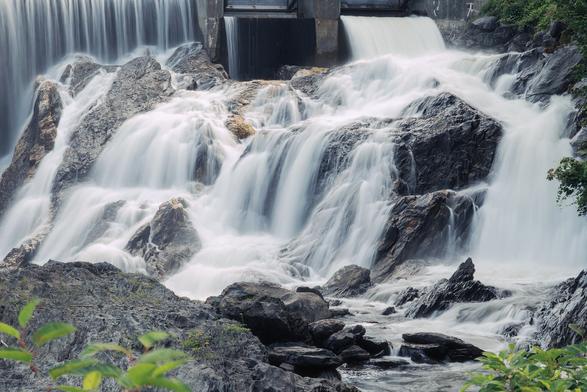 A cascading waterfall flows over rocky ledges, creating a serene and powerful scene with lush greenery in the foreground.