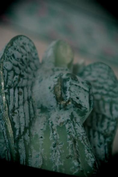 A close up, looking upward at an ornament in the shape of an angel. The ornament is designed to resemble an aged weatherworn statue, there is no face.  The in camera effect emulates a plastic camera with soft focus, teal green colour cast, and vignetting.
