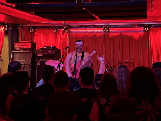 Guitarist and drummer on a small stage, guitarist wearing a yellow sleeveless anime shirt and a mock banzai headband.