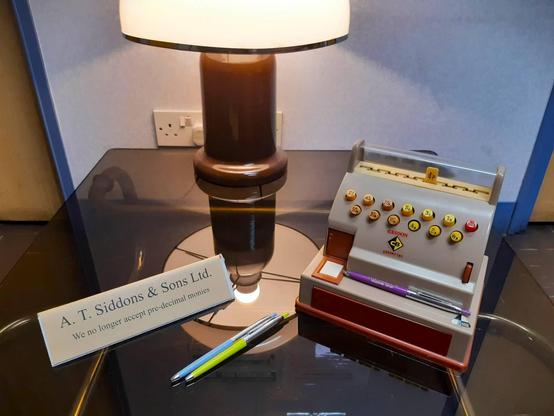 Photo of my 1970 toy cash till sitting on a reception room table with some period correct ballpoint pens. On the table is also a sign that states "We no longer accept pre-decimal monies".

In the background is a 1970s table lamp.

Not in the picture, but inside the till is a selection of 1970s play money which includes 1/2 pennies!