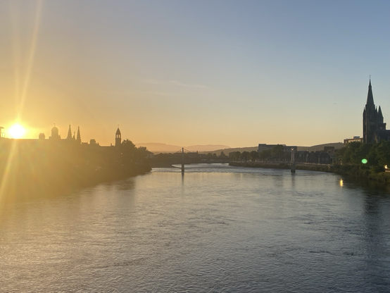 Wide river with an orange setting sun on the left hand side of the picture. Hazy hills in the distance and interesting buildings poking into the orange-blue skyline either side of the water.