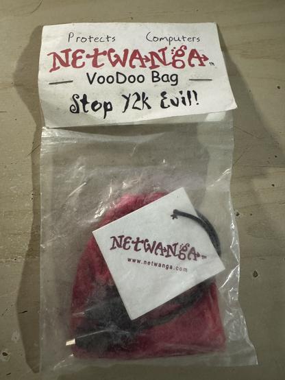 A plastic bag in which is a small velvet bag with two small alligator clips attached to it. There is also a square tag which says “Netwanga” on it. The bag has a cardboard level saying “Protects computers. Netwanga Voodoo Bag. Stop Y2K Evil!