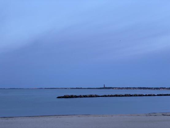 Von unten nach oben ein Streifen Strand, Wasser ohne Wellen, ein steinerner Wellenbrecher, dahinter wieder Wasser ohne Wellen und irgendwann das gegenüberliegende Ufer der Förde mit dem Ehrenmal-Turm in Laboe. Darüber ein sich verdunkelnder Himmel mit verschieden Nuancen an Blau und Grau.