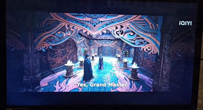 A demon lair clad in red and black. The black carpet has a swirling red pattern that might be phoenixes or dragons - it's hard to tell. The black wall panels has red tree like carvings, and to topp it off the hall is lined with dark red piedestals holding glowing chrystals. An ominous mist is floating above the floor in the background.