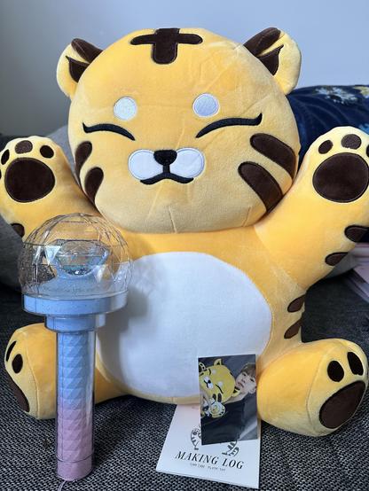 A large, smiling yellow plush toy resembling a tiger (TamTam) sits prominently in the image. In front of it is Seventeen light stick with a diamond-shaped top, along with a photo card displaying Hoshi holding a similar tiger plush.