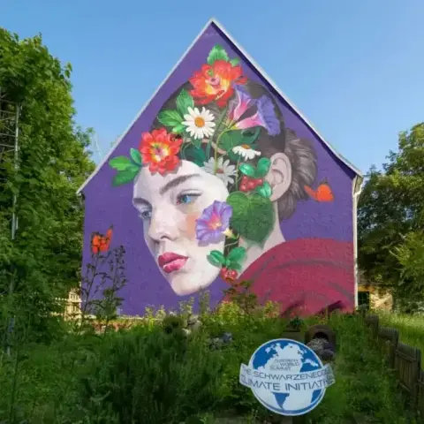 A vibrant mural by street artist Natalia Rak in Asparn an der Zaya, Austria, painted for the Schwarzenegger Climate Initiative. The artwork depicts a woman's serene face in profile with her hair transforming into a lush bouquet of flowers and leaves, including daisies, poppies, and morning glories. Set against a vivid purple background on the side of a house, the mural blends environmental themes with striking realism and surreal beauty. A sign for the Schwarzenegger Climate Initiative is visible in the foreground, surrounded by tall grass and greenery.