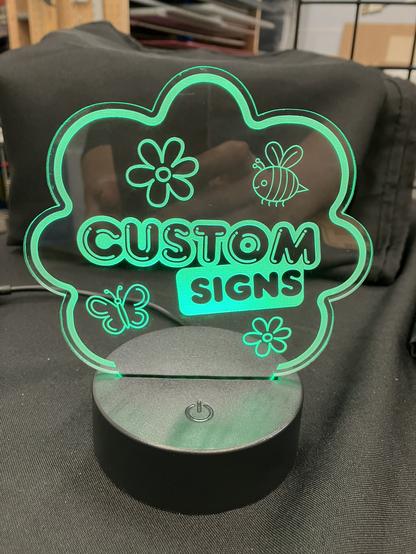 An edge-lit sign with black base is in the shape of a cloud, has a bee, a butterfly, and Flowers on it, and says custom signs in fun bubbly font.