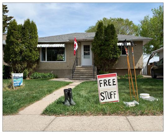 The image shows the front of a house, which appears to be a bungalow with a textured facade and a Canadian flag hanging by the door. On the lawn there are several items apparently offered for free, as indicated by a "FREE STUFF" sign. The items include a pair of black boots, a floor scale, and some walking poles standing vertically next to the sign. There is also a "PROTECT OUR WATER" yard sign suggesting environmental advocacy. The scene is set in a residential area with trees and a walkway leading to the house.