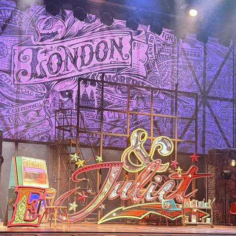 The image features a theatrical stage set for a performance. The backdrop displays the word "LONDON" in stylized lettering, accompanied by various illustrations related to the city. In the foreground, brightly colored props include the names "& Juliet" in vibrant lights