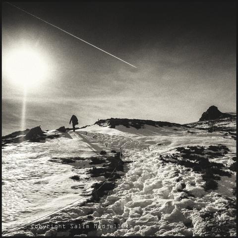 Photographie en noir et blanc du Sancy, prise en contre jour avec une trainée de vol d'un avion qui part de la diagonale gauche et indiquant un sommet sur la droite de la photographie. Une personne en contre jour sur la gauche en ascension..
Black and white photograph of the Sancy, taken against the light with a trail of aircraft flight starting from the left diagonal and pointing to a summit on the right of the photograph. A person in backlight on the left, climbing...