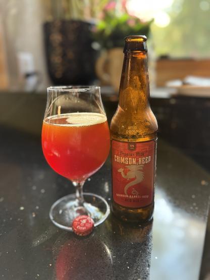 A tall snifter glass of ruby red ale next to a brown bottle with a deep red label and a silver dragon in silhouette