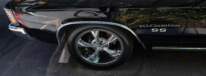 A side view of the front of a gloss-black Chevy El Camino SS, showing the nose and the shiny chrome of its bumper, model badges, wheel, and other trim.  This photo is in the wide 65:24 (or XPan) ratio.