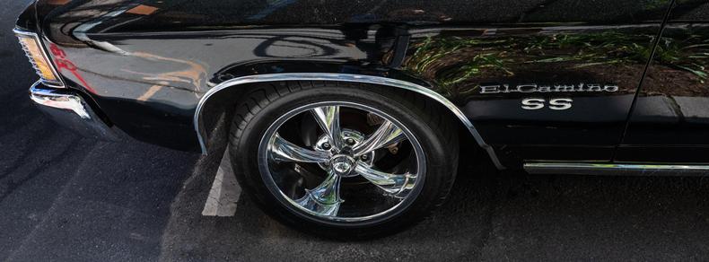 A side view of the front of a gloss-black Chevy El Camino SS, showing the nose and the shiny chrome of its bumper, model badges, wheel, and other trim.  This photo is in the wide 65:24 (or XPan) ratio.