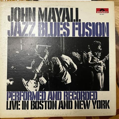 The cover of John Mayall's Jazz Blues Fusion album. The title above the photo, and more text below that says "performed and recorded live in Boston and New York," are rendered in a stencil font. The photograph is rectangular black and white, showing Mayalll's band playing, showing a black man (Freddy Robinson) playing electric guitar, anodht erplayer with his back to us, and John Mayall at the far right, barely in the picture, playing guitar and with a harmonica in a holder near his mouth. Other players are barely visible in the background.