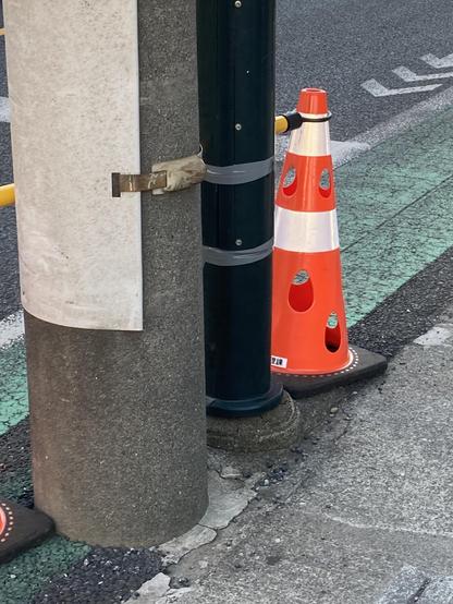 The image shows a gray concrete pole with a poster attached, adjacent to a dark green pole secured with gray tape. A bright orange traffic cone is positioned nearby, with a yellow strap connecting it to the green pole. A bike lane is visible on the ground.