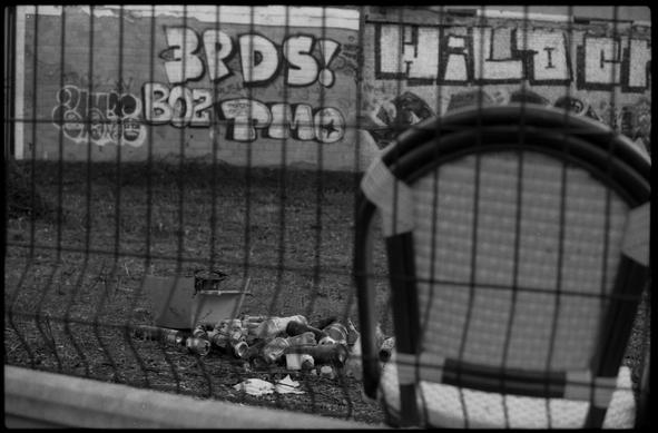 Black and white photography of some trash behind a fence. In the foreground, also behind a fence a chair. in the background some graffitis on a wall.