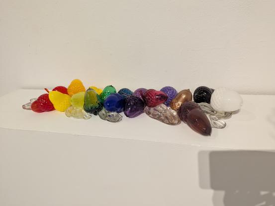 A bunch of acorns and seeds rendered slightly larger than life in a rainbow of bright glass colors on a white pedestal