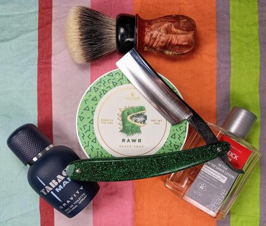 (Le Chat Mistral description)

The image showcases a collection of grooming items arranged on a striped background. At the centre, there's a round tin of shaving soap labelled "RAWR" with a dinosaur graphic. To the left, there's a black bottle of "Tabac Man" eau de toilette. A shaving brush with a wooden handle and white bristles is positioned at the top. To the right, there's a bottle of "Speick" aftershave lotion. A green glittery safety razor lies diagonally across the image. Lastly, a silver straight razor is placed near the shaving soap tin. The items are neatly arranged, suggesting a well-organised grooming routine.